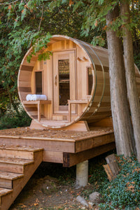 Dundalk Leisurecraft Canadian Timber Tranquility Barrel Sauna with Front Porch, placed outside near trees in a backyard facing left